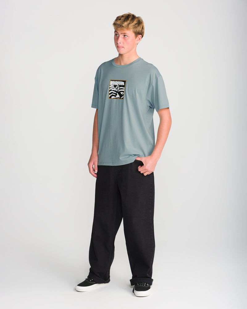 A person wears a grey Volcom Men’s Hand Held Short Sleeve Tee with a rectangular chest graphic featuring a black and white abstract pattern, paired with black pants and black sneakers.