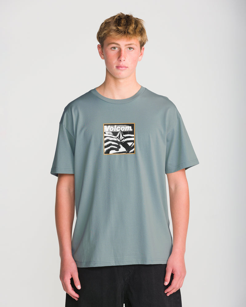 A person wearing a grey Volcom Men’s Hand Held Short Sleeve Tee, featuring a graphic on the chest of a hand holding a stone shape with text.