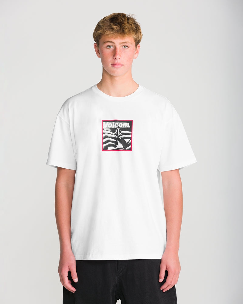 A young man wears a heavyweight, loose-fit white Men’s Hand Held Short Sleeve Tee by Volcom, featuring a black and white graphic design with the brand name in a red rectangular frame on the chest.