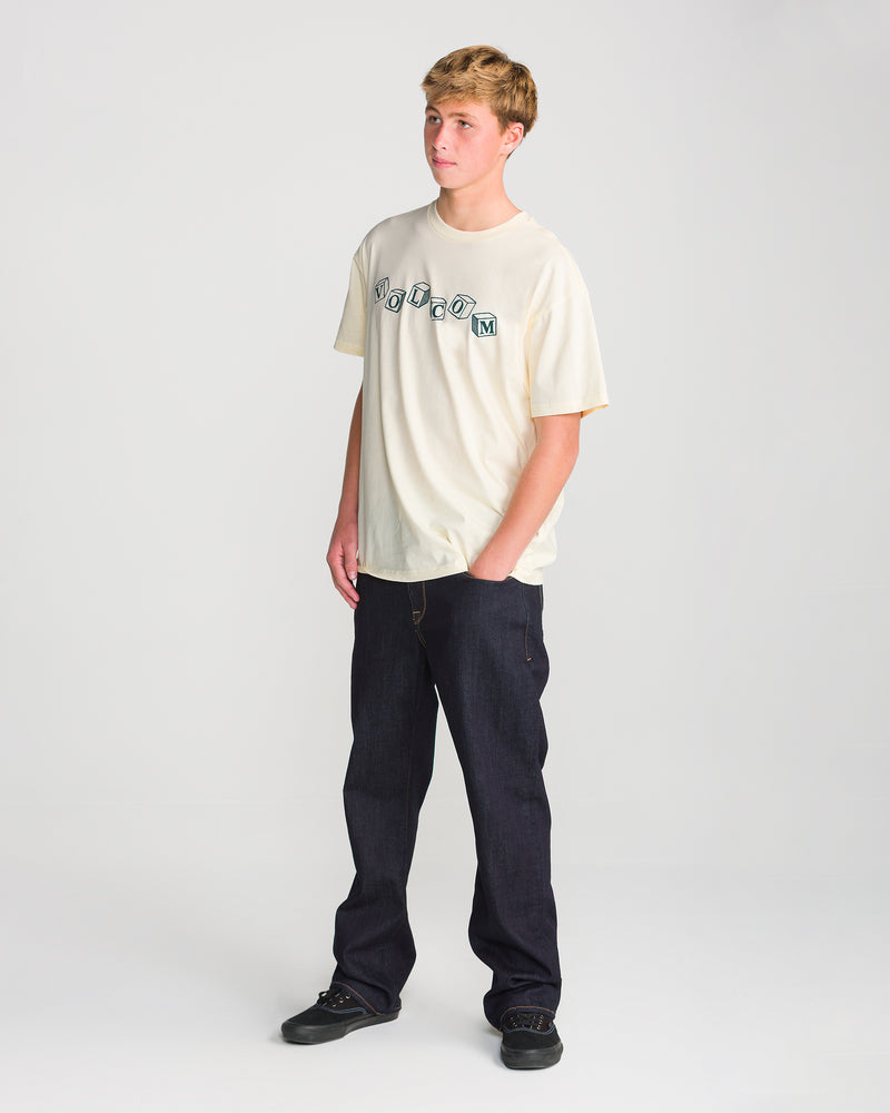 A person wearing a loose fit, off-white Volcom Men’s Last Laugh Short Sleeve Tee with block lettering across the chest, dark jeans, and black sneakers.
