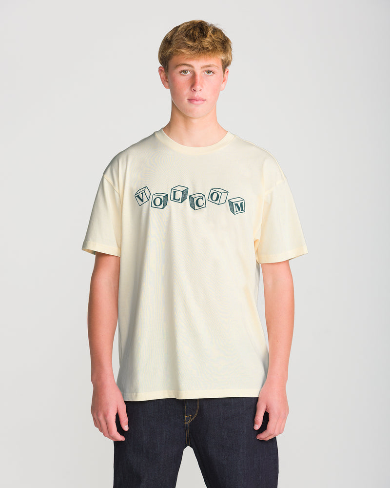 A young man wearing an off-white, loose-fit Men’s Last Laugh Short Sleeve Tee with the word VOLCOM printed in a block-letter style that resembles alphabet cubes.