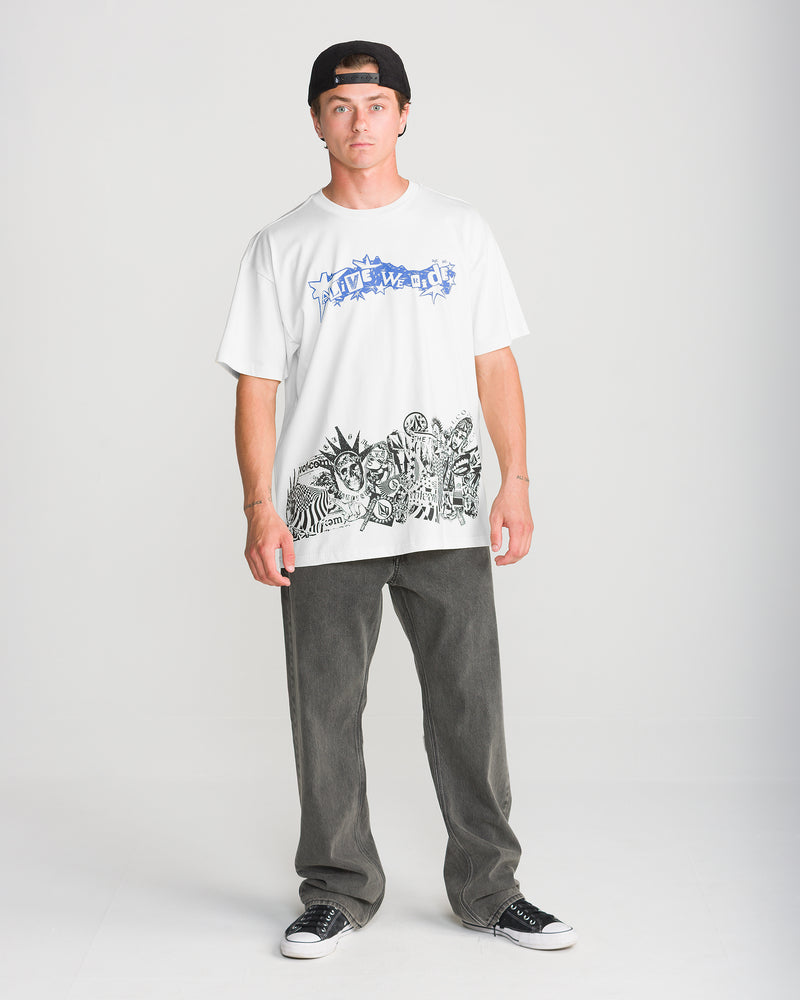 A man wearing a Volcom Men’s Alive We Ride Short Sleeve Tee with blue text across the chest and black graphic prints along the hem, paired with grey jeans and black sneakers.