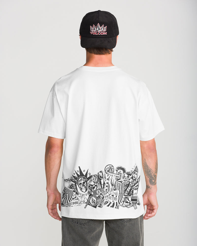A person wears a Volcom Men’s Alive We Ride Short Sleeve Tee, featuring a black baseball cap and a white t-shirt with a black-and-white graphic print of skulls and geometric patterns across the lower back.