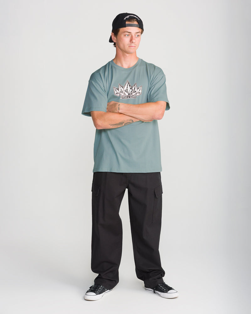 A man wearing a green Volcom Men’s Tooms Short Sleeve Tee, black cargo pants, and a baseball cap stands with his arms crossed.