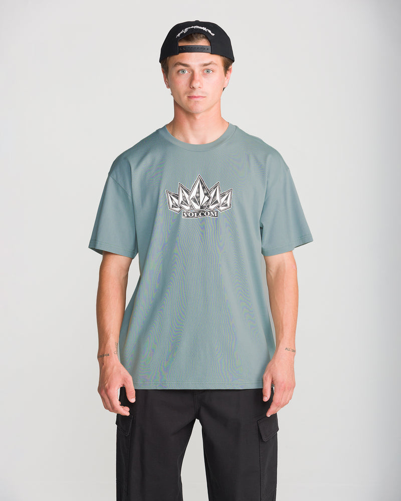 A man wearing a sage green Volcom Men’s Tooms Short Sleeve Tee with a white graphic logo on the chest and a black baseball cap.