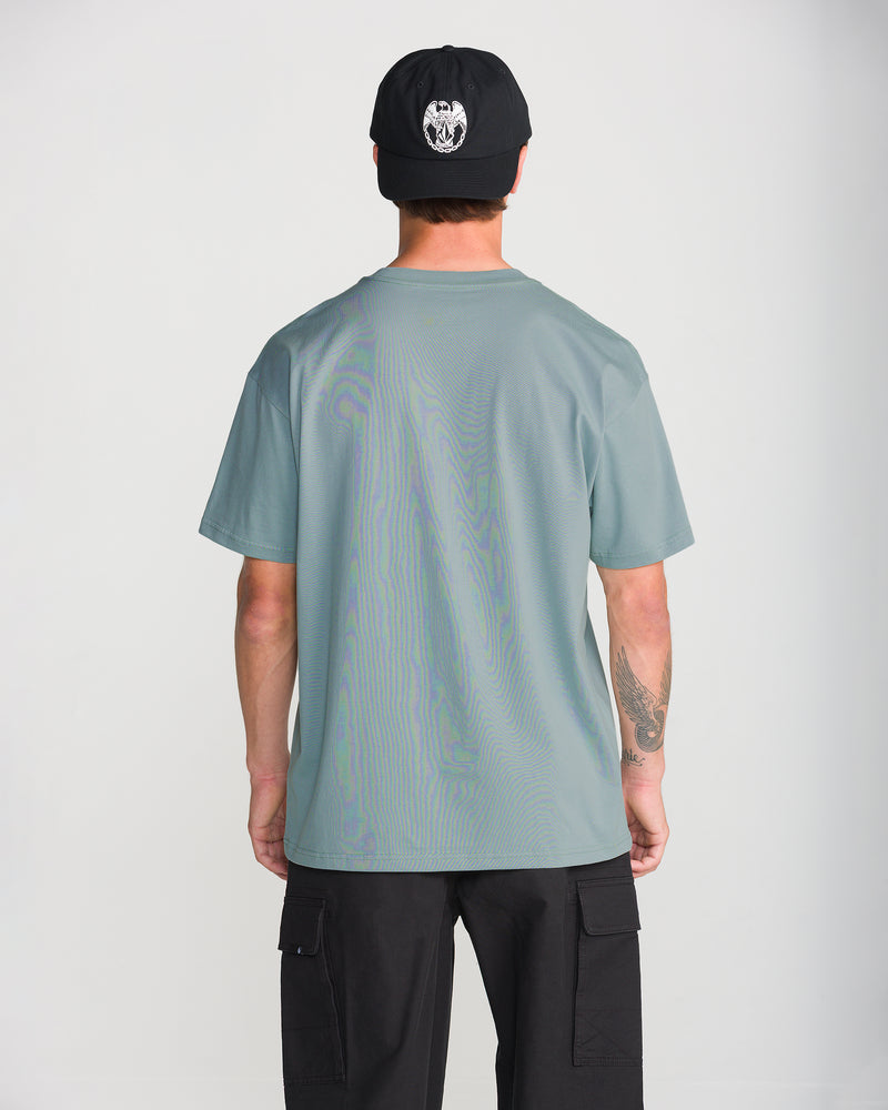 Back view of a person wearing a loose fit sage green heavyweight cotton Volcom Men’s Tooms Short Sleeve Tee, black cargo shorts, and a black baseball cap.