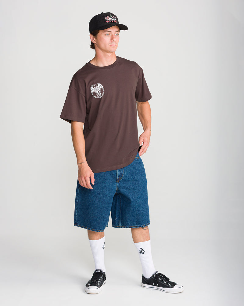 A man wearing a brown Volcom Men’s Linked Up Short Sleeve Tee with a chest graphic, blue denim shorts, white socks, and black sneakers.