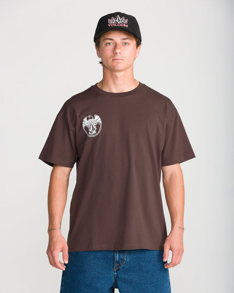 A man wears a black baseball cap and a brown Volcom Men’s Linked Up Short Sleeve Tee featuring a white eagle graphic on the chest.
