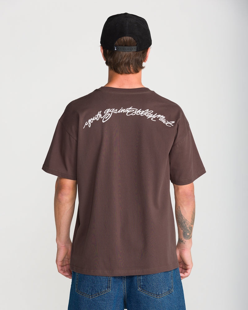 Back view of a person wearing a brown, loose-fit Volcom Men’s Linked Up Short Sleeve Tee with youth against establishment written in white script across the upper back.