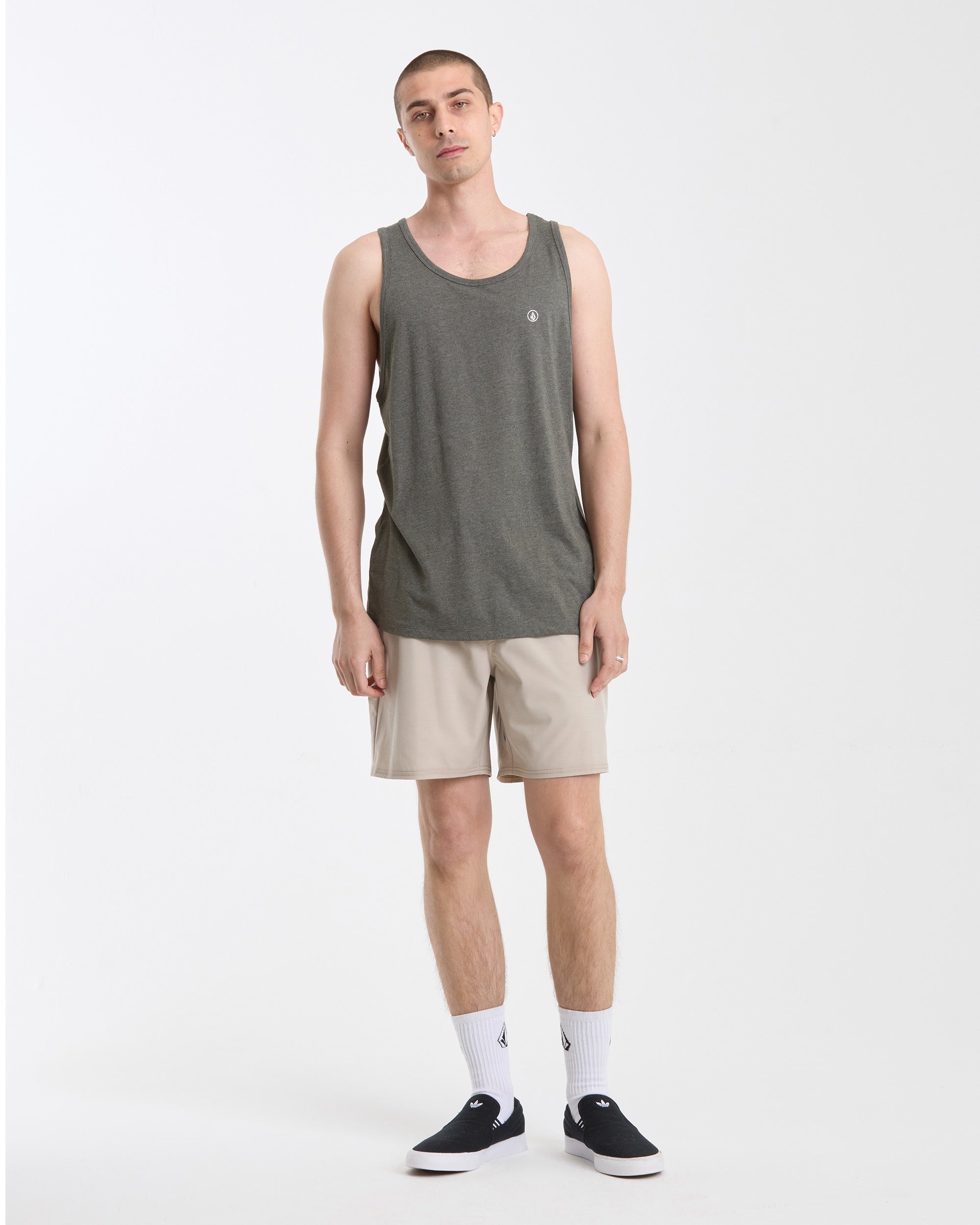 A man stands against a plain white background in the Volcom Men's Solid Heather Tank, paired with beige shorts, white crew socks, and black slip-on shoes. His arms are relaxed by his sides as he faces the camera with a neutral expression.