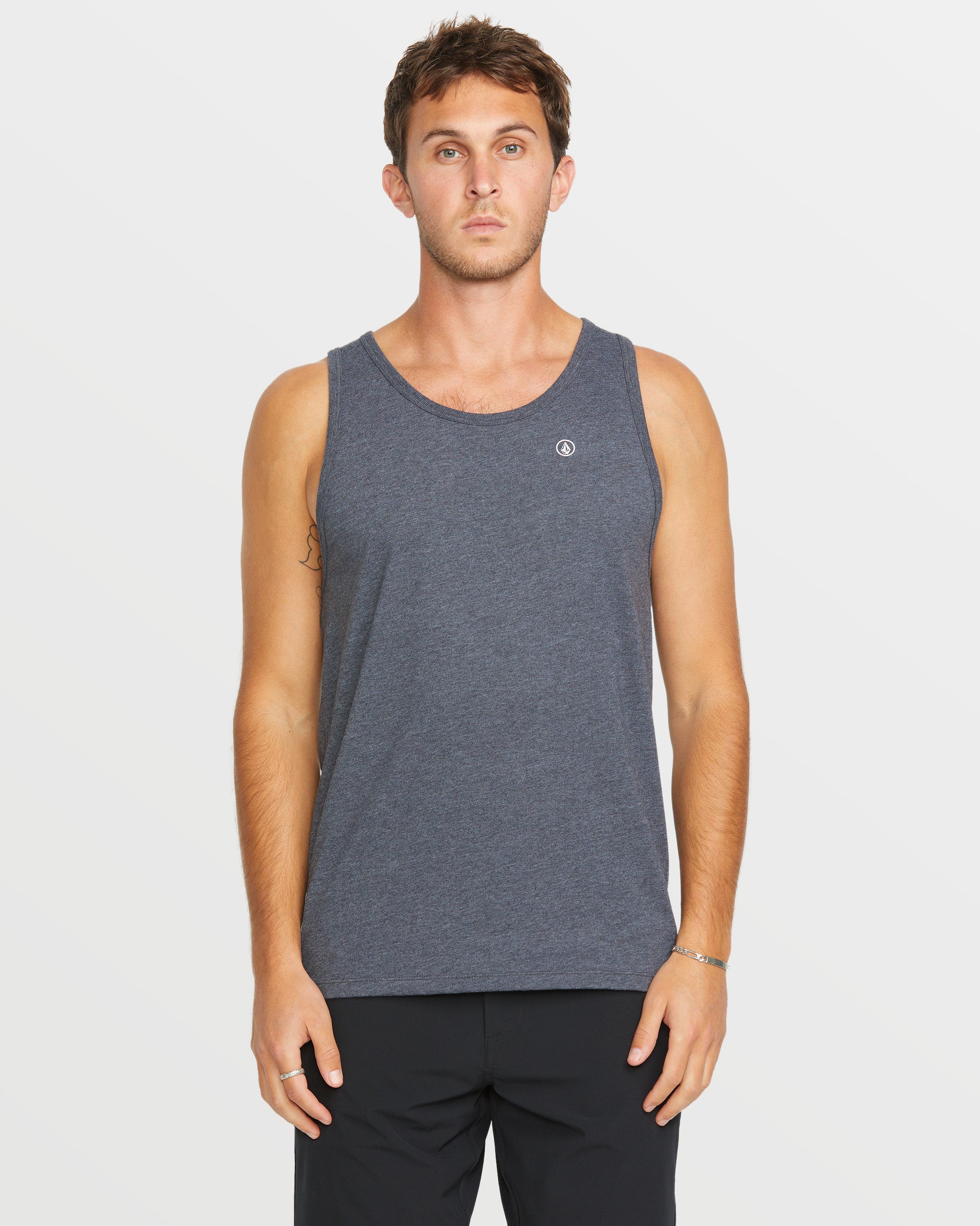 A man stands facing forward against a plain white background, wearing the Volcom Men's Solid Heather Tank in dark gray paired with black pants.