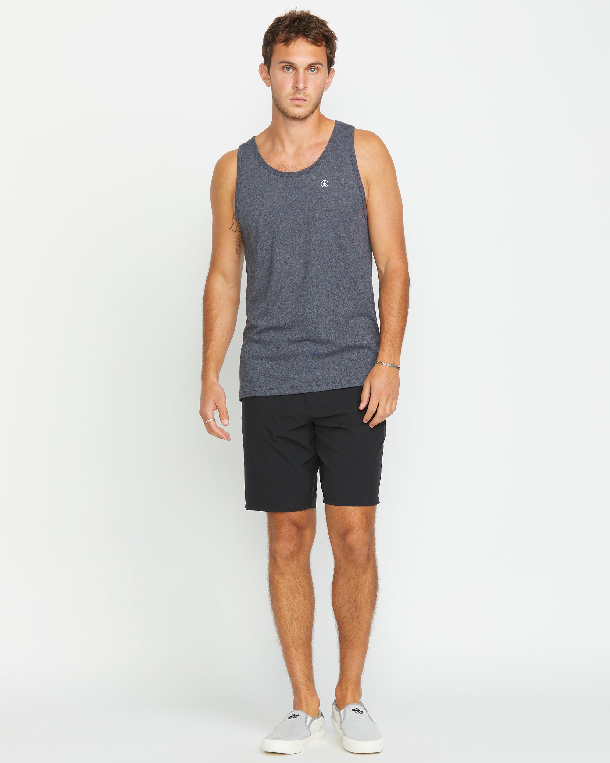 A man stands facing forward against a plain white background, wearing the Volcom Men's Solid Heather Tank in dark gray, paired with black shorts and light gray slip-on shoes.