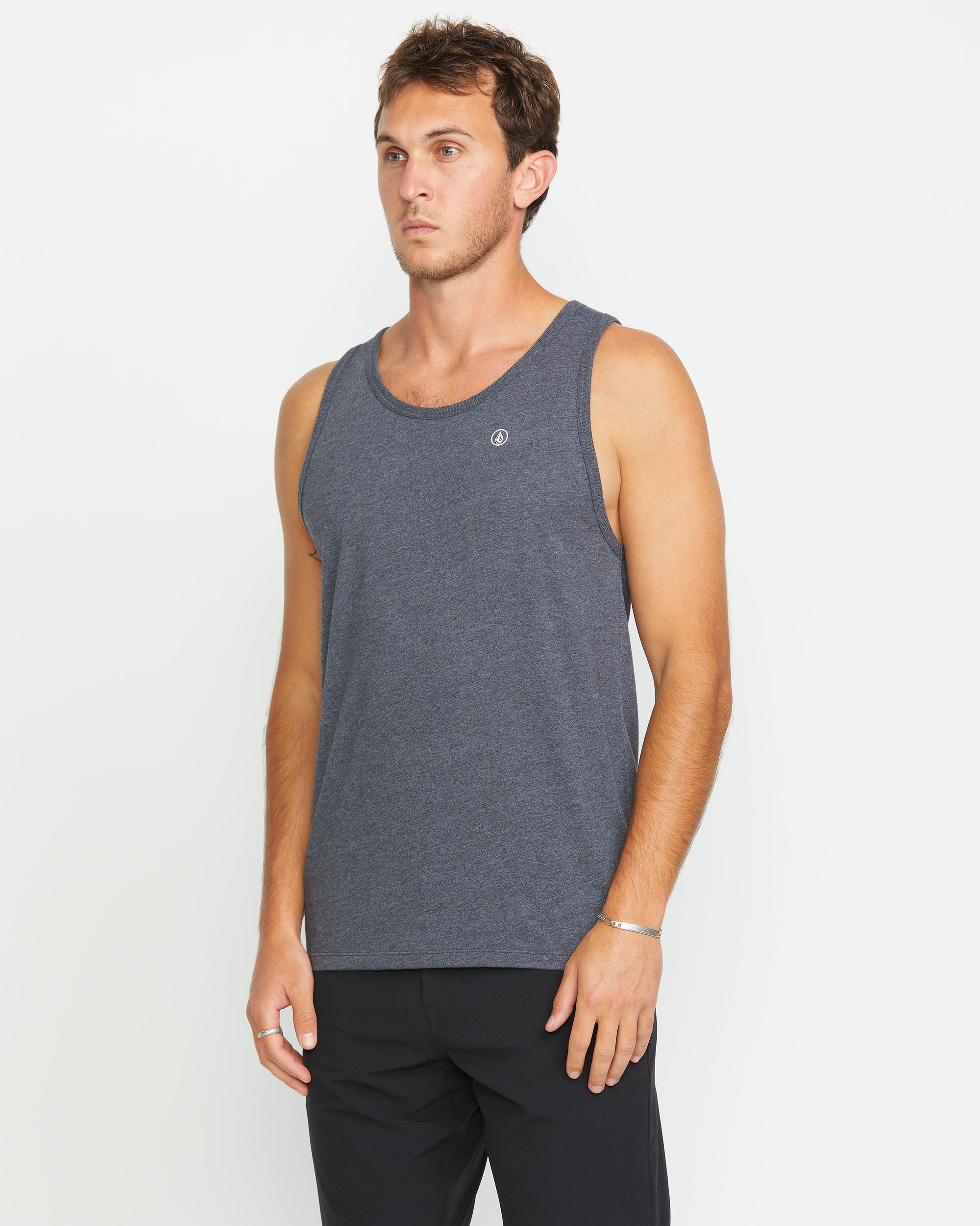 A man wearing the Volcom Men's Solid Heather Tank in dark gray and black pants stands against a plain white background, gazing slightly to the side with a neutral expression.