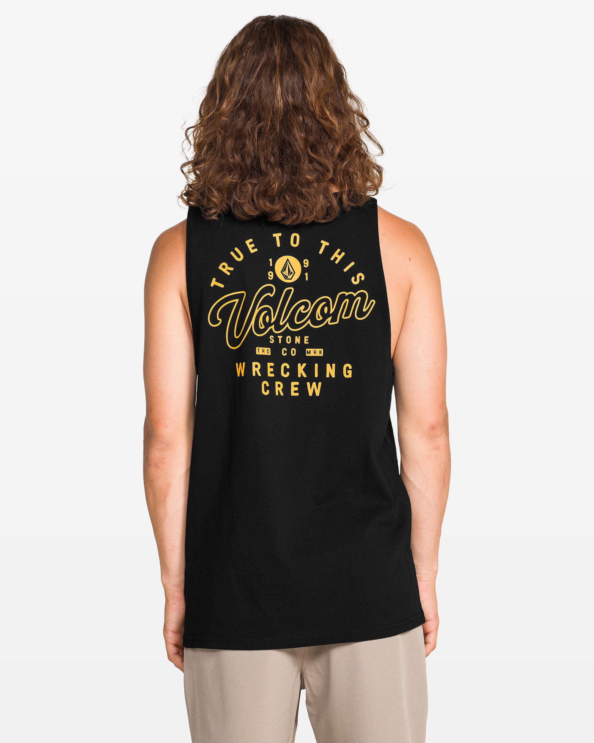 A person with long curly hair faces away from the camera, wearing a classic fit black Volcom Men's Wrecking Tank made of 100% cotton, featuring yellow "True To This Volcom Stone Co Wrecking Crew" text on the back.