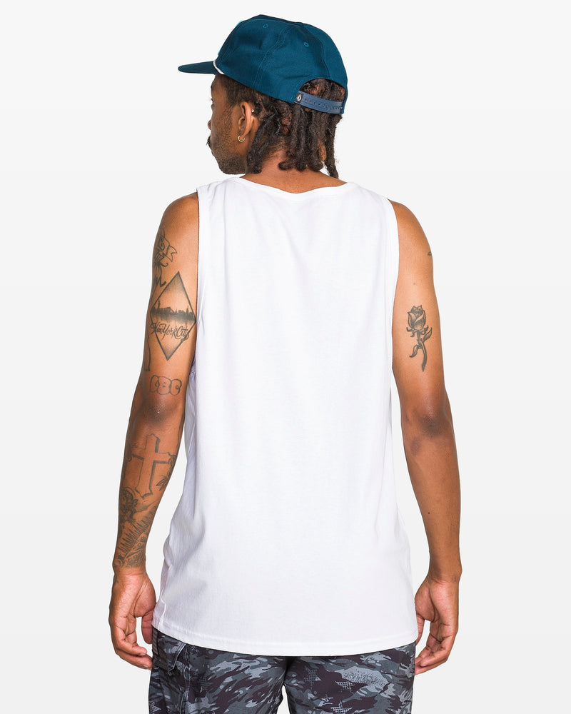 A tattooed individual stands with their back to the camera against a white backdrop, wearing the Volcom Men's Serve Stoked Tank from the Froth of July collection, patterned shorts, and a backwards dark blue cap.