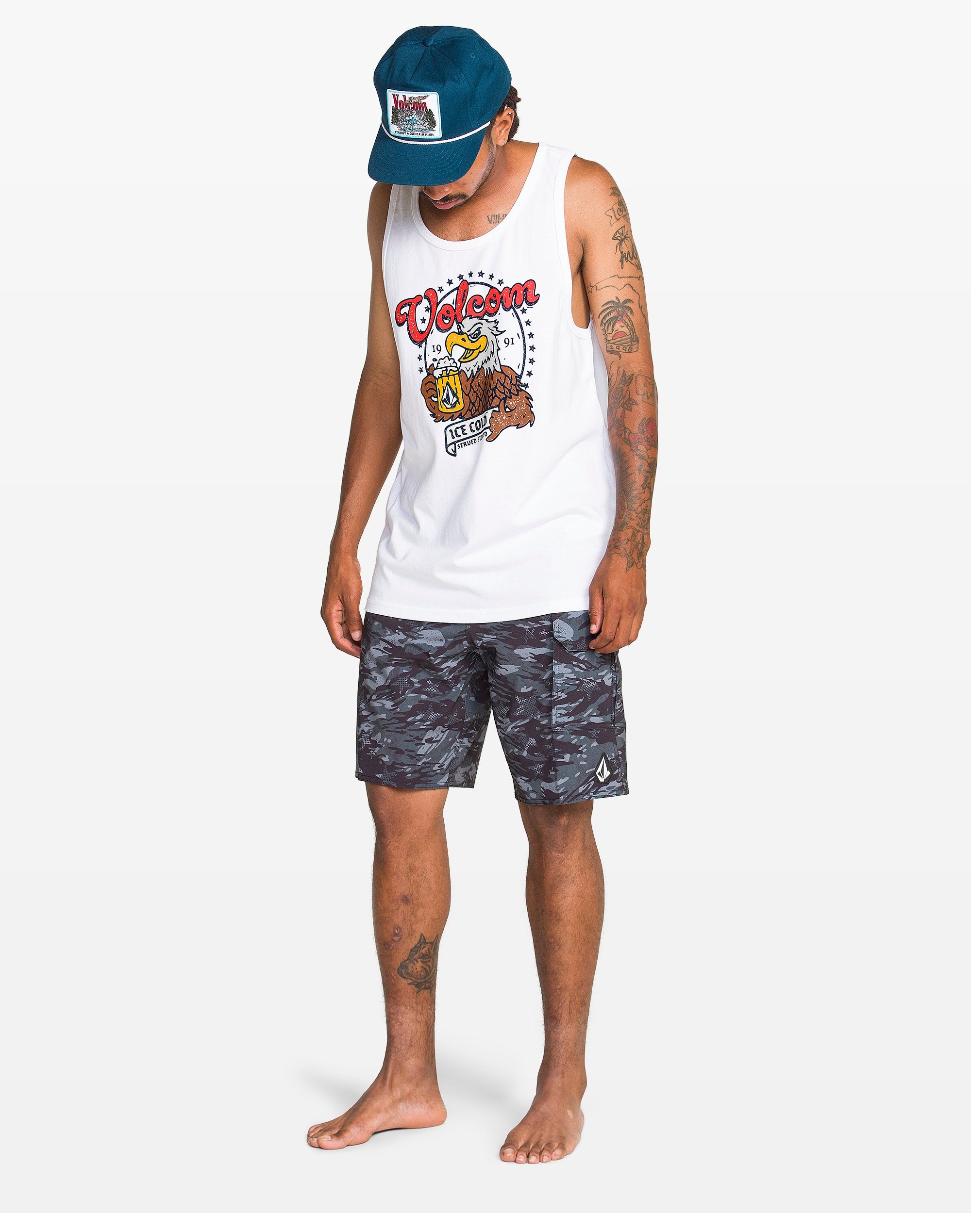 A man in a blue cap, Volcom Men's Serve Stoked Tank, and black camo shorts stands barefoot looking down against a plain white background.