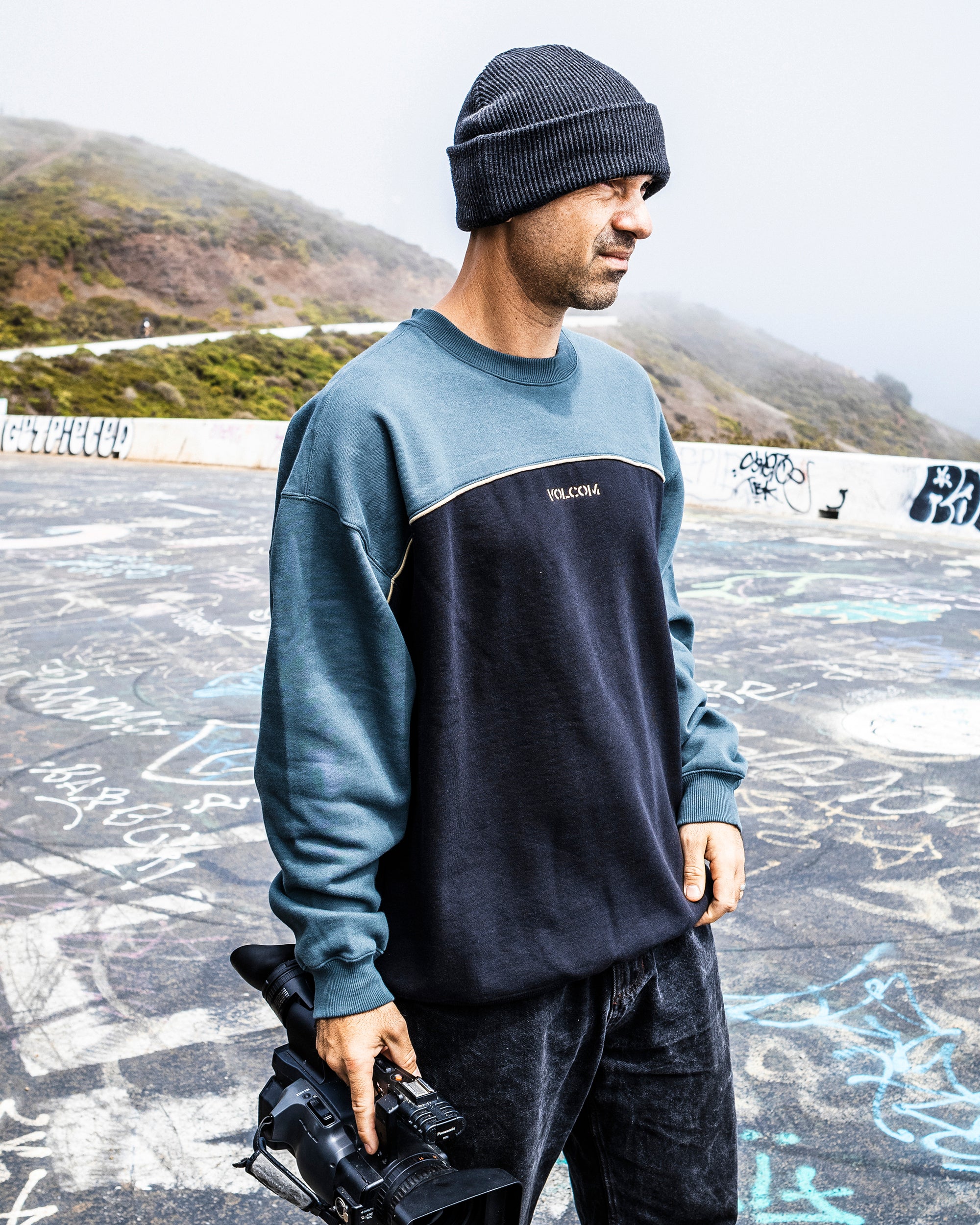 A man in a Volcom Men's 2K Stone Crew beanie stands outdoors on graffiti-covered concrete, holding a video camera, with misty hills behind him.