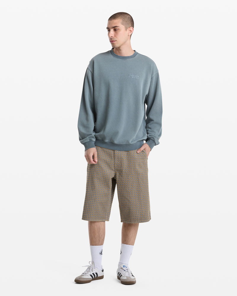 A person stands against a plain white background wearing the Volcom Men's Smoothie Crew sweatshirt, patterned knee-length shorts, white crew socks, and gray Adidas sneakers, looking to the side with one hand in their pocket.