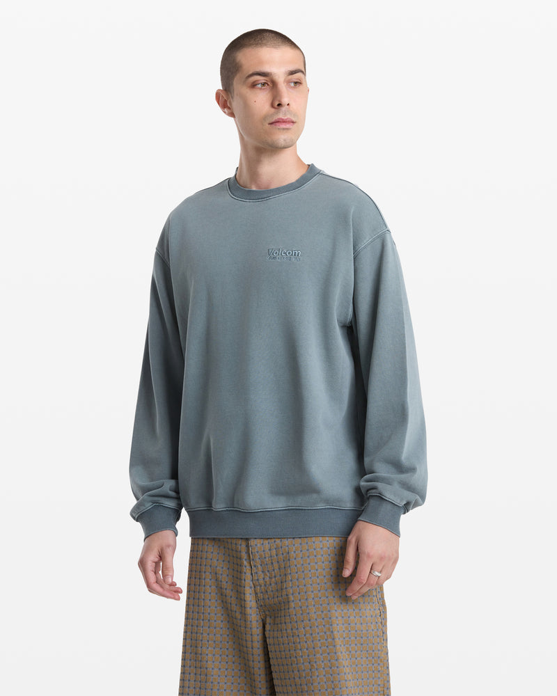 A man with short hair wears the Volcom Men's Smoothie Crew, a loose-fitting faded blue sweatshirt with Volcom embroidery, paired with patterned brown shorts. He stands against a plain white background, looking slightly to the side.