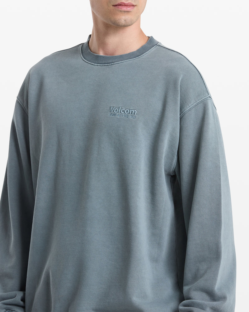A person wears the Volcom Men's Smoothie Crew, a loose-fitting, faded blue crewneck sweatshirt with an embroidered Volcom logo on the chest. The background is plain white and the person's head is partially visible.