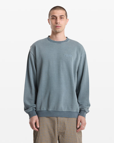 A person with short hair wears a loose-fitting, faded blue Volcom Men's Smoothie Crew sweatshirt and patterned brown pants, standing against a plain white background.