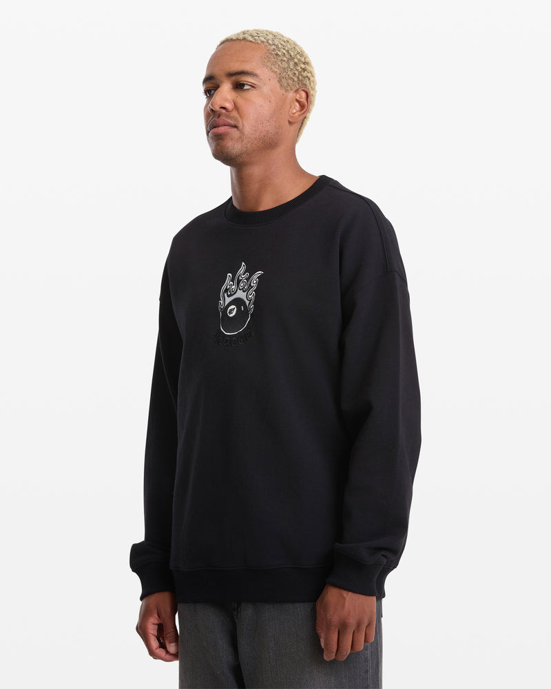 A man with short blond hair wears the Volcom Men's Featured Artist Seth Conboy Crew, a loose black fleece crewneck with a small white flaming vinyl graphic on the chest. He stands against a plain white background, looking slightly left.