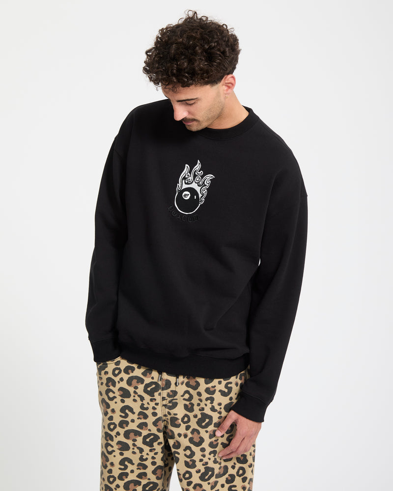 A man with curly hair wears the Volcom Men's Featured Artist Seth Conboy Crew, a black fleece sweatshirt with a flaming yin-yang graphic, paired with beige leopard print pants, standing against a plain white background.