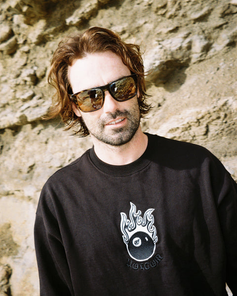 A man with wavy brown hair and a beard wears large sunglasses and the Volcom Men's Featured Artist Seth Conboy Crew sweatshirt, standing in front of a rocky wall.