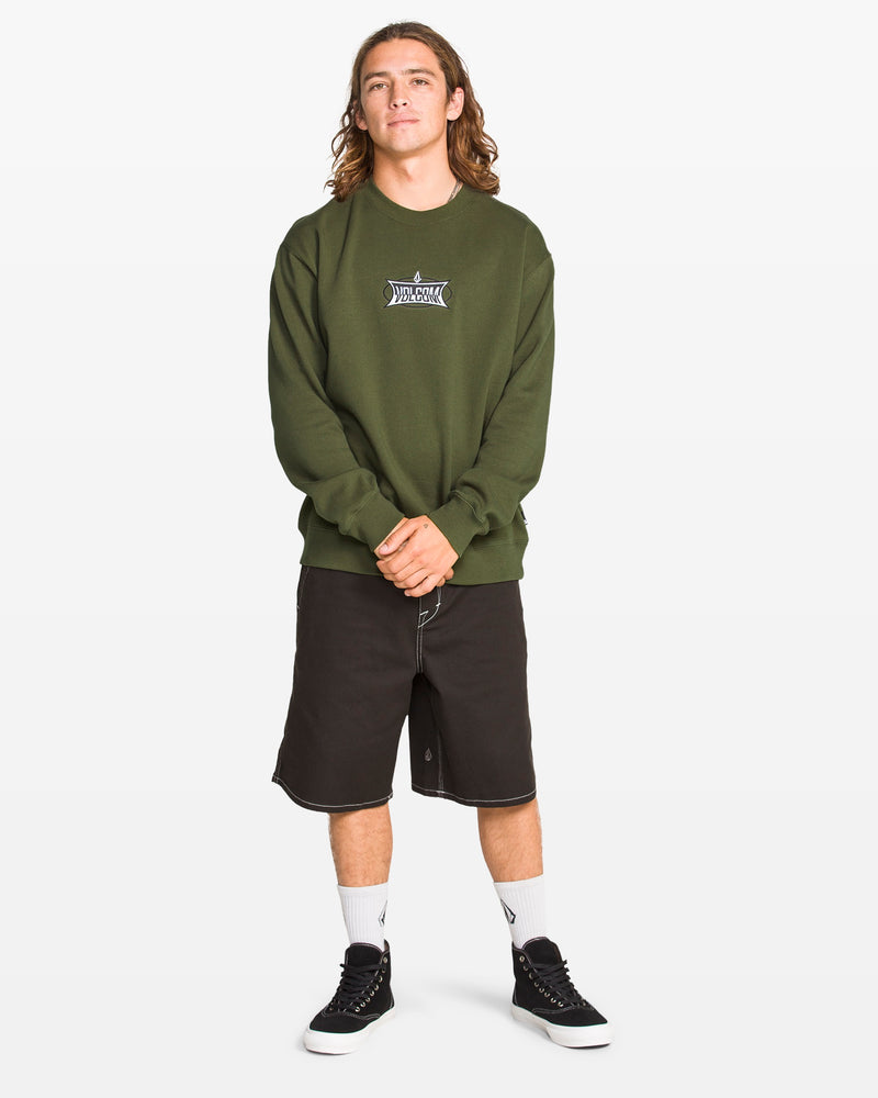 A person with long hair stands facing forward, wearing a Volcom Men's Stone Cruiser Crew in Squadron Green, made from recycled polyester, paired with black knee-length shorts, white socks, and black sneakers against a plain white background.