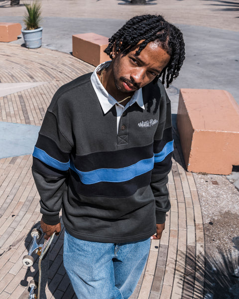 A man with short dreadlocks stands outdoors holding a skateboard and looking up at the camera, dressed in the Volcom Men’s Scratch Stone Fleece Polo paired with jeans.