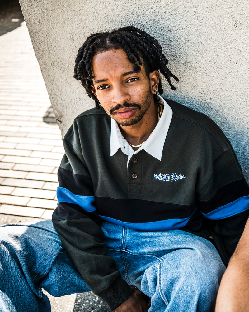 A person with short dreadlocks and a trimmed beard sits against a wall, wearing the Volcom Men’s Scratch Stone Fleece Polo—a dark green and blue colorblocked collared shirt made with REPREVE recycled polyester. Paired with light blue jeans, they look into the camera with a neutral expression, highlighting the boxy fit and modern style of this sustainable fleece polo.