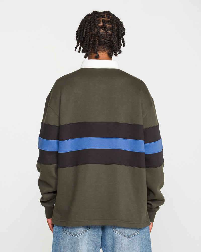 A person with short twists stands facing away, wearing the Volcom Men's Scratch Stone Fleece Polo in a boxy fit olive green with black and blue horizontal stripes, crafted from recycled polyester fleece, paired with light blue jeans against a plain white background.