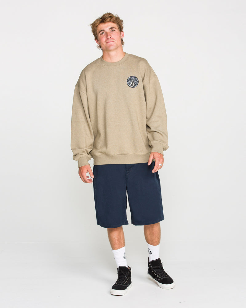 A young man stands against a plain white background, wearing the Volcom Men's Dirty Work Crew—an easygoing beige (khaki) brushed-back fleece sweatshirt with a small circular logo—paired with navy blue shorts, white socks, and black sneakers. The loose fit of the Volcom sweatshirt enhances his relaxed expression as he looks at the camera.