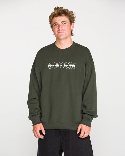 A person with short, tousled hair wears the Volcom Men's Dirty Work Crew— a green, loose fit fleece crew sweatshirt crafted from recycled polyester—paired with black pants and standing against a plain light background. The sweatshirt features white text for added style and offers both comfort and sustainability.