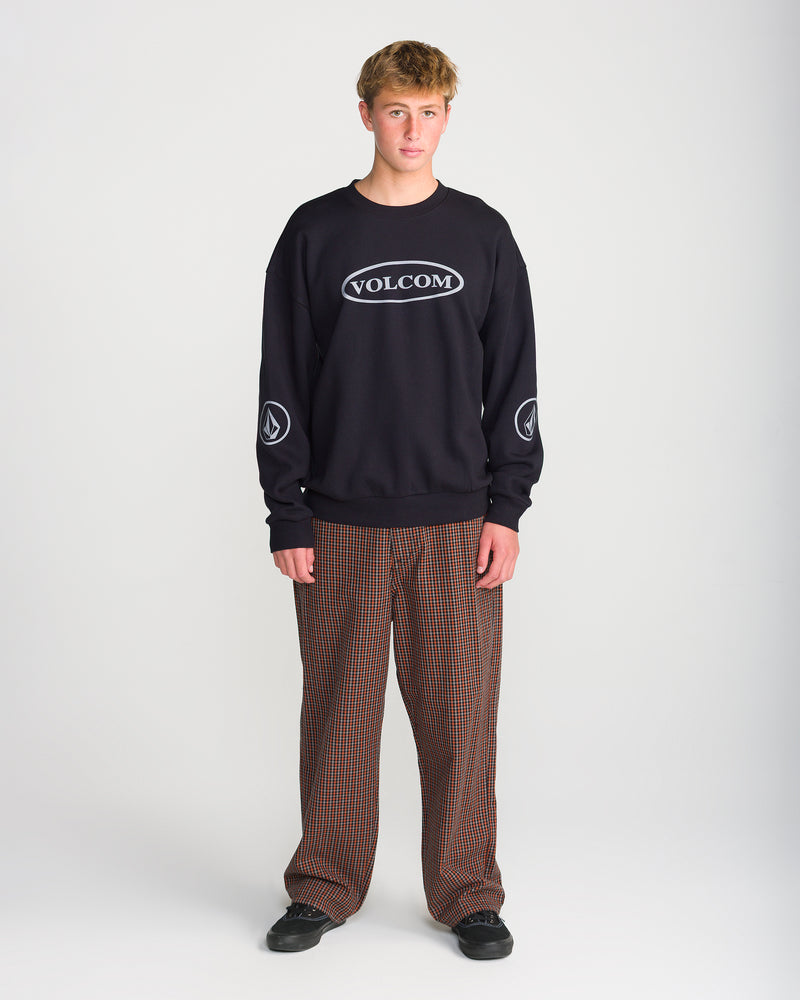 A person wearing a loose fit black Men’s Stone Op Crew with white Volcom graphics on the chest and sleeves, paired with brown plaid trousers and black sneakers.