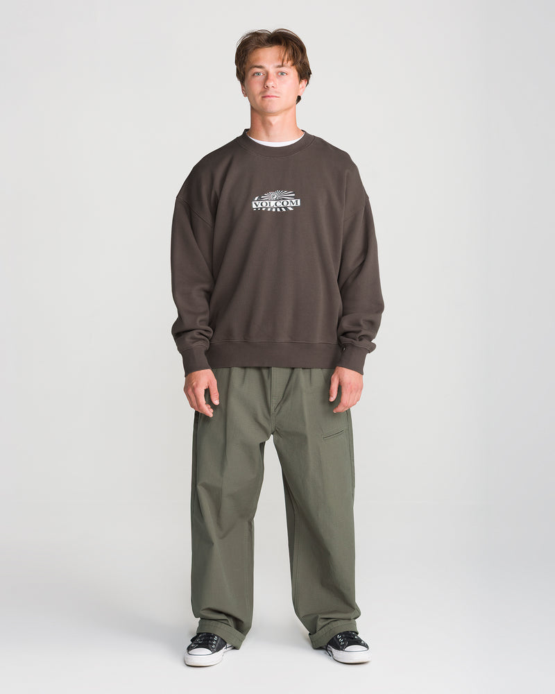 A person wearing a dark brown vintage wash Volcom Men’s Stone Op Crew in a boxy fit with a graphic logo on the chest, olive green trousers, and black sneakers.