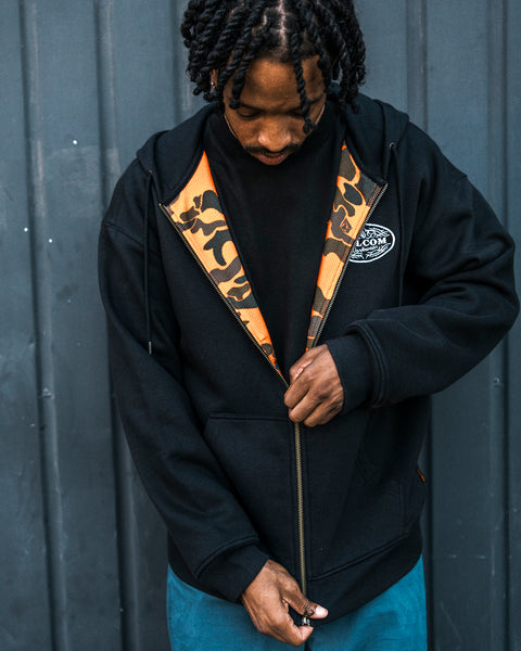 A person with braided hair looks down while zipping up the Volcom Men’s On The Hunt Workwear Zip, a black fleece featuring a camouflage-patterned lining made from recycled polyester, as they stand in front of a dark metal wall.