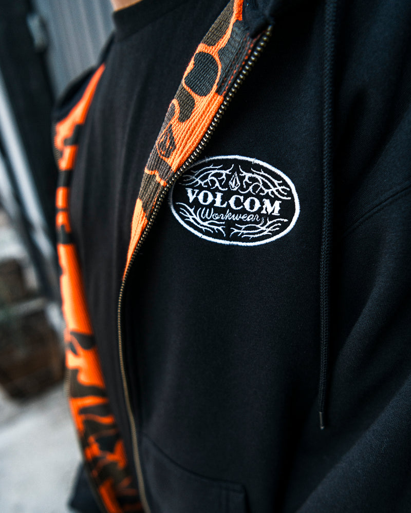 A person wearing the Volcom Men’s On The Hunt Workwear Zip hoodie, featuring a black exterior and an orange patterned cotton waffle thermal lining. Partially unzipped to reveal the chest logo, this fleece is crafted from recycled polyester for durability, making it stand out against the blurred background.