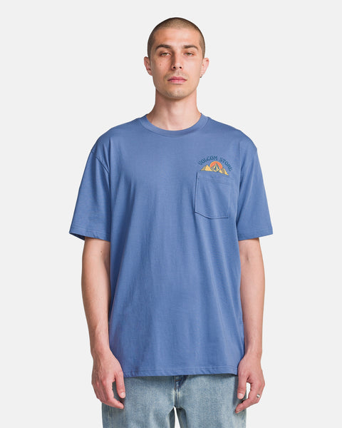 A man wears the Volcom Men's Great Divide Short Sleeve Tee—blue ring spun cotton with a front pocket and small mountain graphic on the left shoulder—paired with light blue jeans, standing against a plain white background.