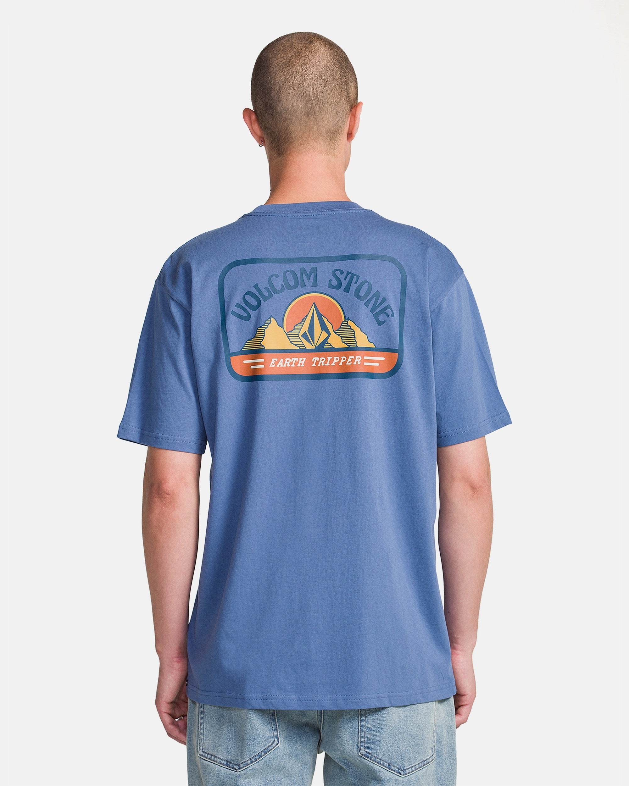 A person faces away from the camera, wearing the Volcom Men's Great Divide Short Sleeve Tee in blue—made of soft ring spun cotton, with a back graphic of mountains, a tent, and the words "VOLCOM STONE" and "EARTH TRIPPER.