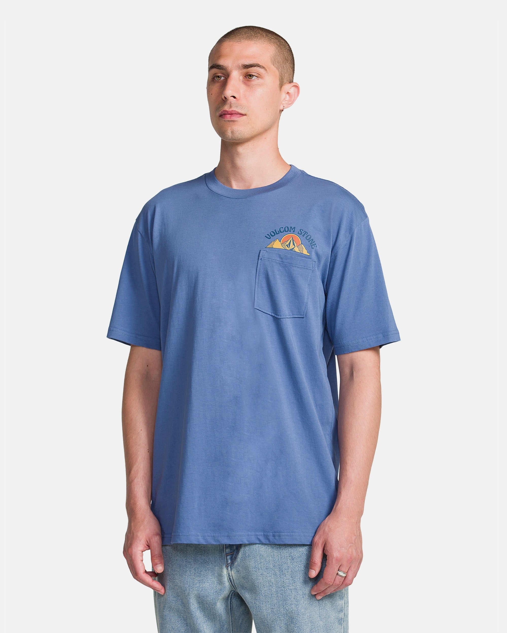A man wearing the Volcom Men's Great Divide Short Sleeve Tee, a relaxed-fit ring spun cotton shirt with a chest pocket and mountain graphic, paired with light blue jeans, stands against a plain white background.