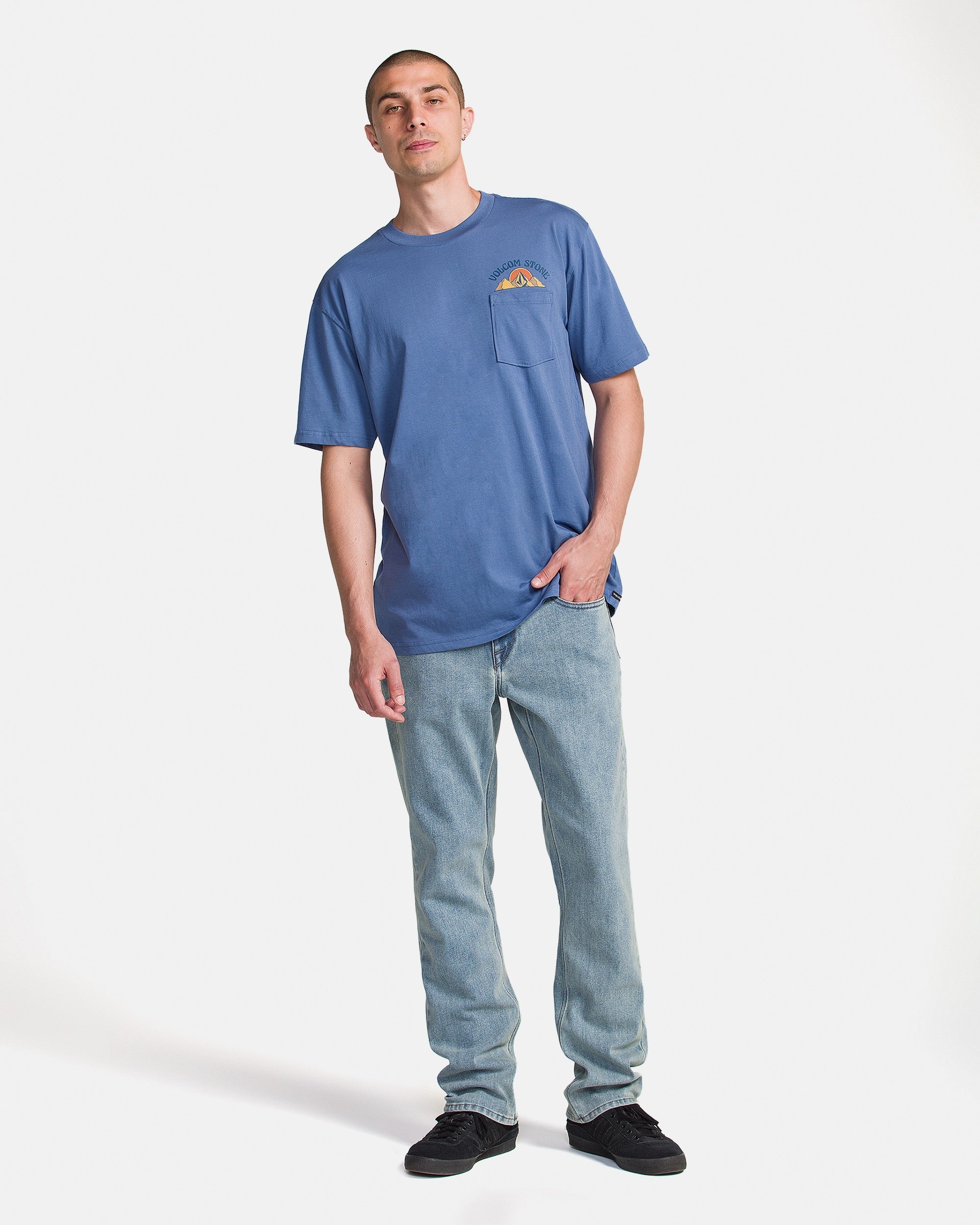 A man with short hair stands against a white background wearing the Volcom Men's Great Divide Short Sleeve Tee, featuring soft ring spun cotton and a chest pocket, paired with light blue jeans and black shoes, hand in pocket.