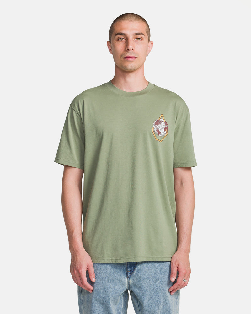 A person faces forward in a Volcom Men's Great Divide Short Sleeve Tee, a soft ring spun cotton shirt in light green with a small, colorful left chest graphic, paired with light blue jeans against a plain white background.