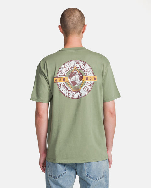 A person with a shaved head is seen from the back wearing light blue jeans and a light green Volcom Men's Great Divide Short Sleeve Tee, made from ring spun cotton with a classic fit and featuring a large circular graphic and text on the back.