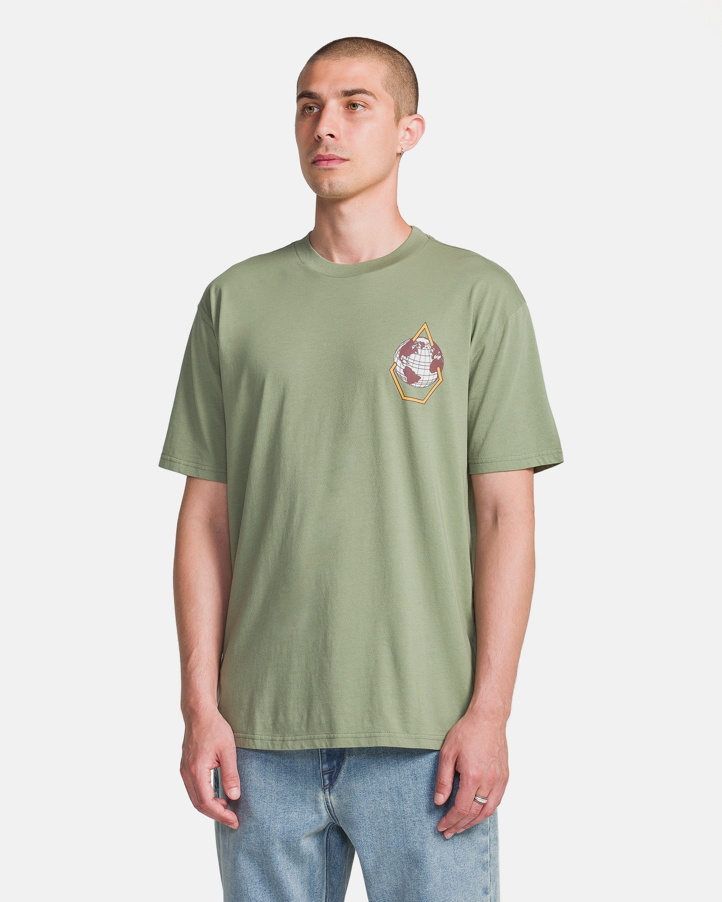 A man wears the Volcom Men's Great Divide Short Sleeve Tee in light olive green, featuring a soft ring spun cotton build and small left chest graphic, paired with light blue jeans against a plain white background.