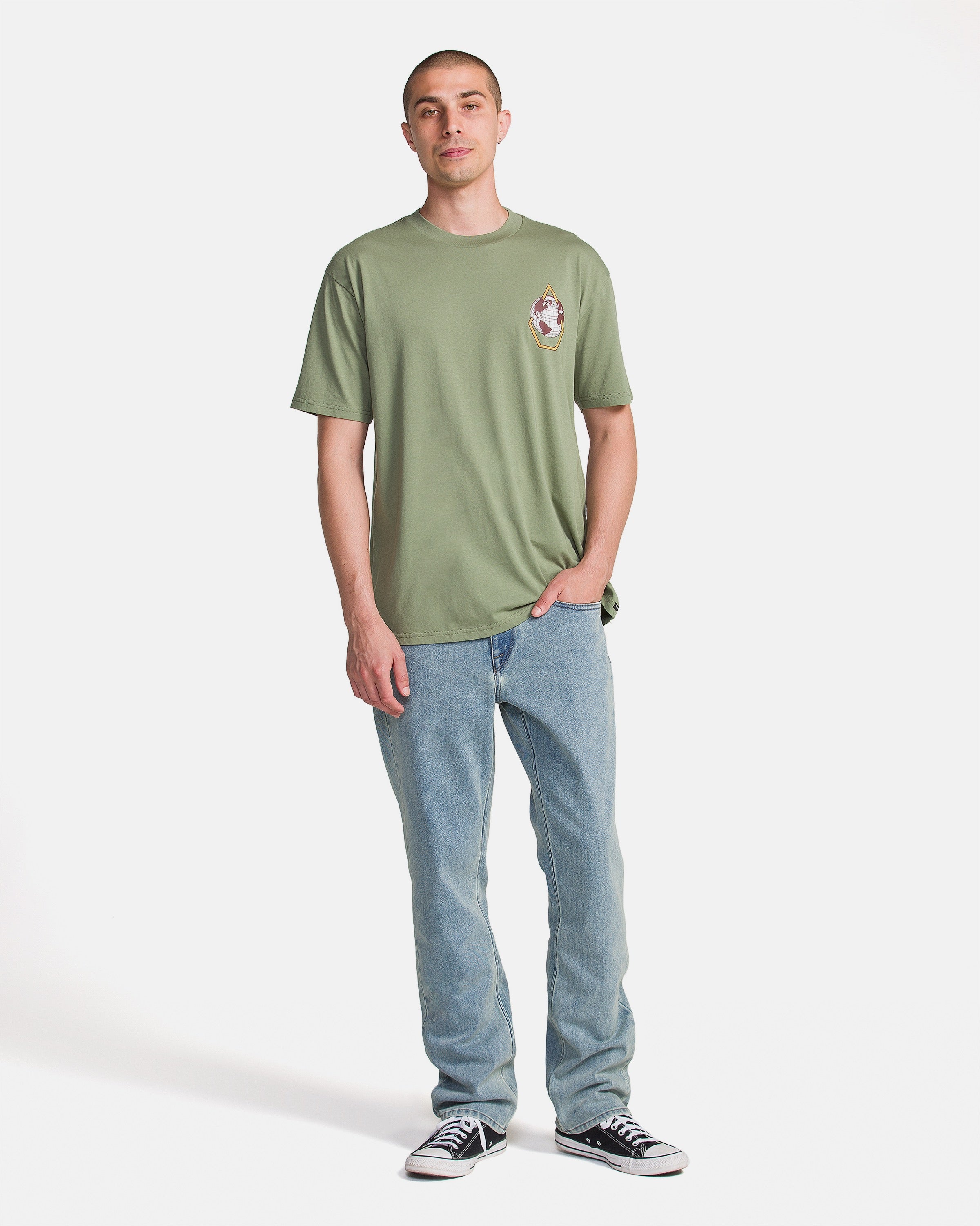 A person stands against a white background, wearing the Volcom Men's Great Divide Short Sleeve Tee in light green with light blue jeans and black-and-white sneakers. One hand is in their pocket as they look at the camera.
