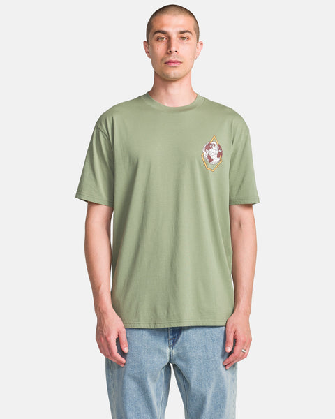 A man wearing the Volcom Men's Globular Short Sleeve Tee in light green with a small left chest graphic and light blue jeans, standing against a plain white background.