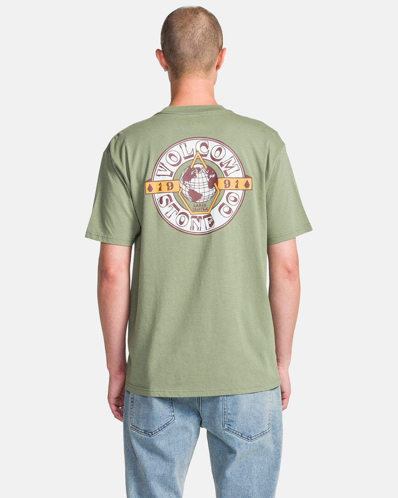 A person in a light green Volcom Men's Globular Short Sleeve Tee with a large circular back graphic, paired with light blue jeans, stands facing away from the camera against a white background.