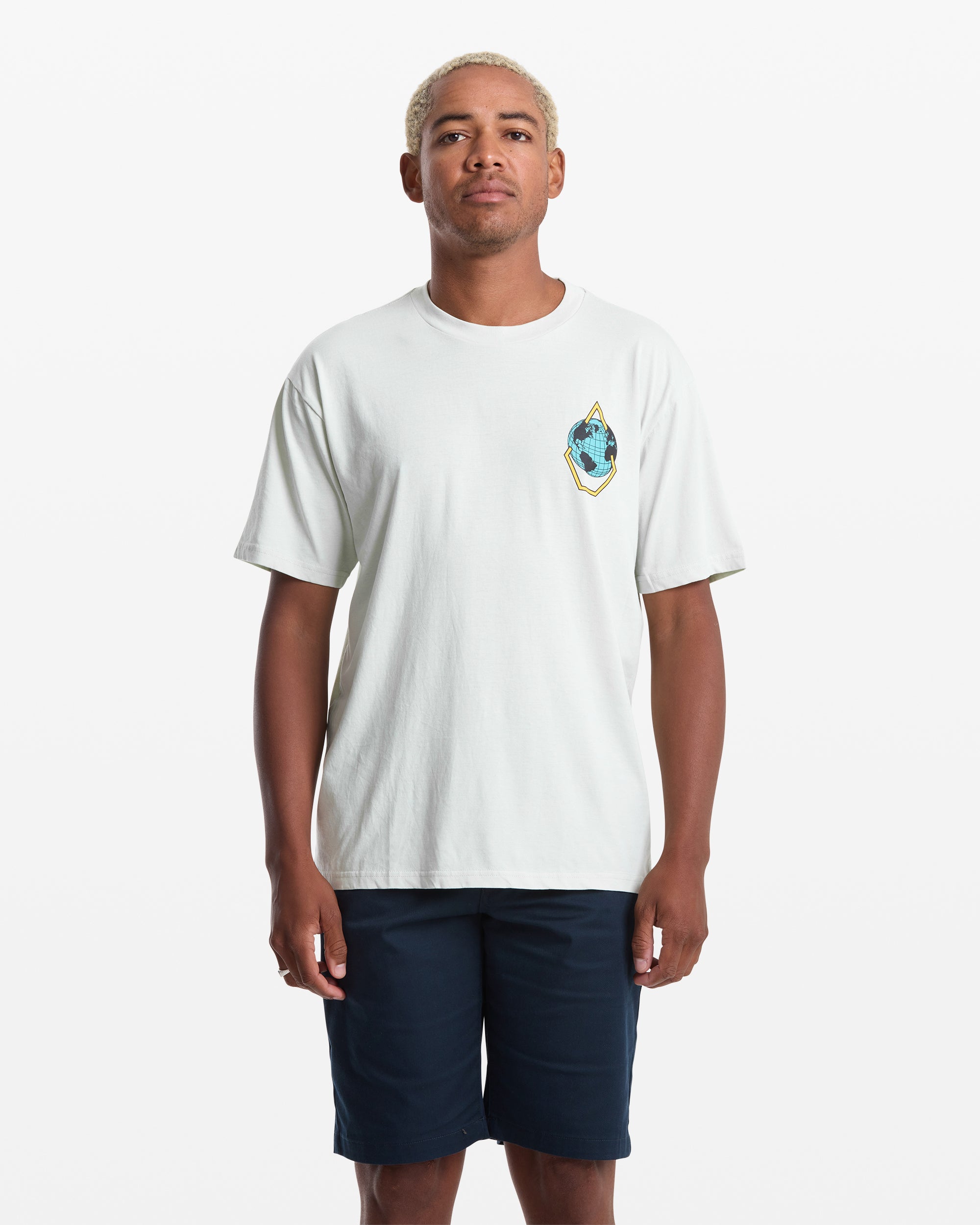 A man with short blond hair wears the Volcom Men's Globular Short Sleeve Tee, a white t-shirt featuring a small colorful chest patch, paired with dark navy shorts against a plain white background.
