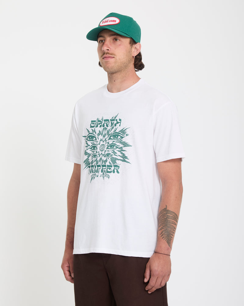 A man wears the Volcom Men's Remote Viewer Short Sleeve Tee—white, featuring a psychedelic face graphic—and brown pants, plus a green and white cap, standing against a plain white background.